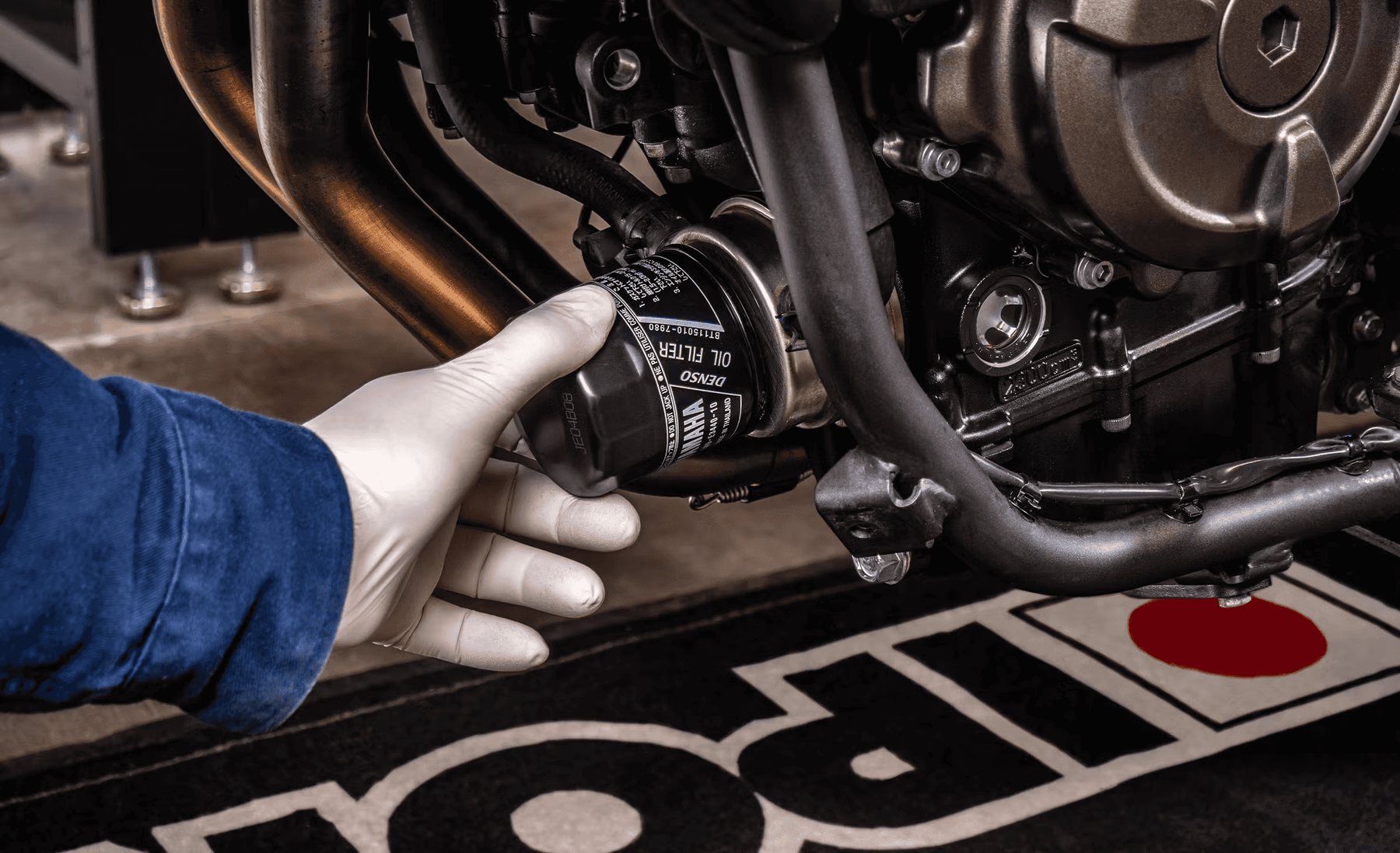 Main gantée présentant un filtre à huile devant un moteur de moto.